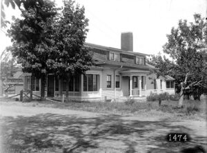 Early 20th-century building with a small rounded front porch and two rectangular dormers on the roof. Two large trees partially block the house from sight. Side-frontal view, circa 1910.