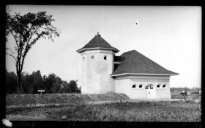 Small early 20th-century square building with steep roofing and small half-circle windows. Attached is a taller round turret with small circular windows on all sides. Frontal view, circa 1908.
